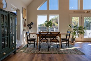 Un comedor con mesa de madera, cuatro sillas, un telescopio, un armario y un ventanal con vistas a árboles.
