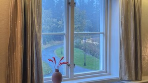 Una ventana con vistas a una carretera y follaje, un jarrón con flores rojas y cortinas.