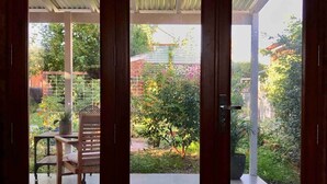 Una habitación con suelo de madera, una mesa con sillas y vistas a un jardín exterior con plantas y una valla.