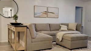 Un salon comprenant un canapé d’angle, un miroir rond, une petite console en bois ornée d’une plante, et deux tableaux encadrés accrochés au mur.