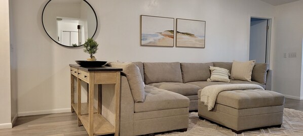 Un salon comprenant un canapé d’angle, un miroir rond, une petite console en bois ornée d’une plante, et deux tableaux encadrés accrochés au mur.