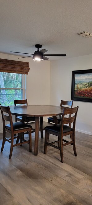 Un coin repas comprenant une table ronde et quatre chaises, un ventilateur de plafond et une fenêtre donnant sur un paysage verdoyant.