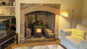 A living room with a stone fireplace, a television on a stand, a sofa with a yellow pillow, and a patterned rug. - A cosy character 18th century cottage with enclosed garden close to Ullswater. (Stainton, near Penrith)