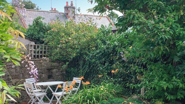 A garden with a stone wall, a white table and chairs, and various plants and flowers. - A cosy character 18th century cottage with enclosed garden close to Ullswater. (Stainton, near Penrith)
