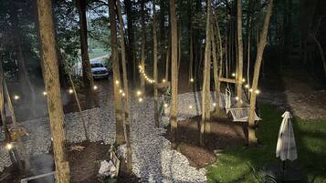 A forested outdoor area with a gravel path, a fire pit, hanging chairs, and string lights.