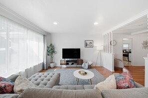 A modern living room with a large sofa, a flat-screen TV, and a dining area with a kitchen in the background.