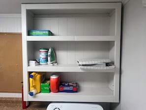 Een witte plank met diverse spullen, waaronder een blik ontsmettingsdoekjes, een strijkijzer, een kussen en schoonmaakmiddelen.