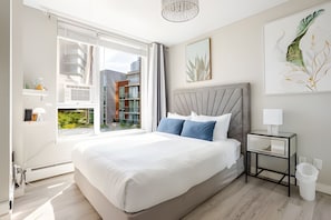A modern bedroom with a large bed, a bedside table, a nightstand, a window, and a view of buildings outside.