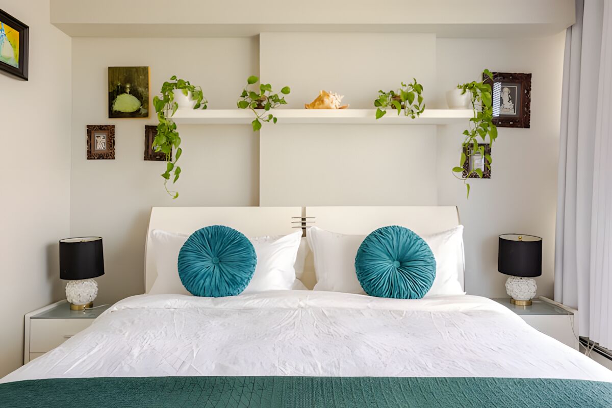 A hotel room with a large bed, two bedside lamps, a white headboard, and a shelf with decorative items and plants.
