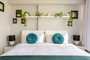 A hotel room with a large bed, two bedside lamps, a white headboard, and a shelf with decorative items and plants. - The Pond - The Full Package (Vancouver)