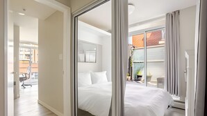 A modern bedroom with a large bed, a sliding glass door leading to a balcony, and a view of a building outside.