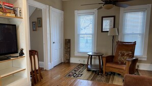 A living room with a television, wooden flooring, a patterned rug, a leather chair, and a ceiling fan. - Renovated Victorian in 'Americas Coolest Small Town' (Hammondsport)