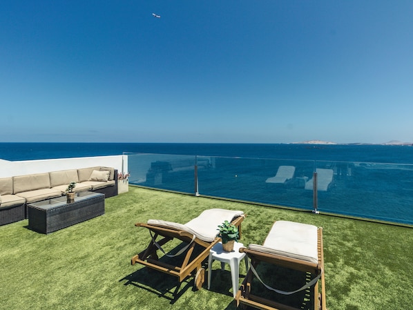 A rooftop terrace with a clear view of the ocean, a glass balustrade, and outdoor furniture including a sofa and two lounge chairs.