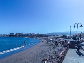 A beach with people swimming and sunbathing, a stone wall, and buildings in the background.