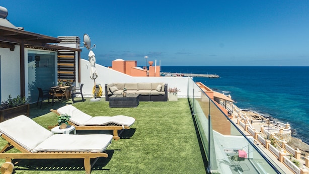 Terraço no terraço com vista clara do mar, mobiliado com cadeiras de descanso e um sofá, e guarda-corpo de vidro com vista para o oceano.