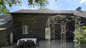 Een stenen gebouw met een metalen pergola in de achtertuin, een tafel met stoelen en een klein raam.