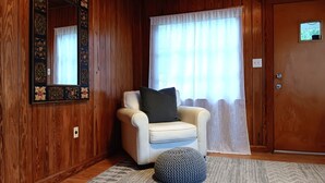 Une pièce aux murs lambrissés, un fauteuil blanc avec un coussin foncé, un pouf rond, un miroir avec un cadre décoratif et une porte dotée d’une petite fenêtre.