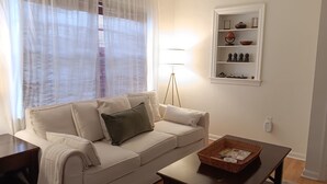 Un salon avec un canapé beige, une petite table, une lampe sur pied et une étagère ornée d’objets décoratifs.