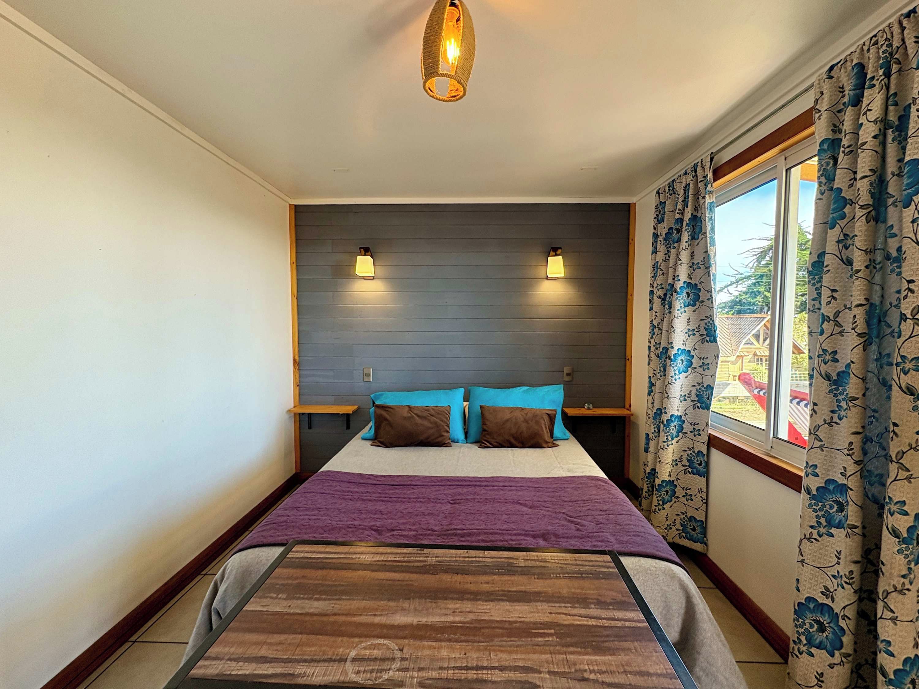 Un dormitorio con cama, cabecera, lámparas de noche, ventana con cortinas y vistas al exterior.