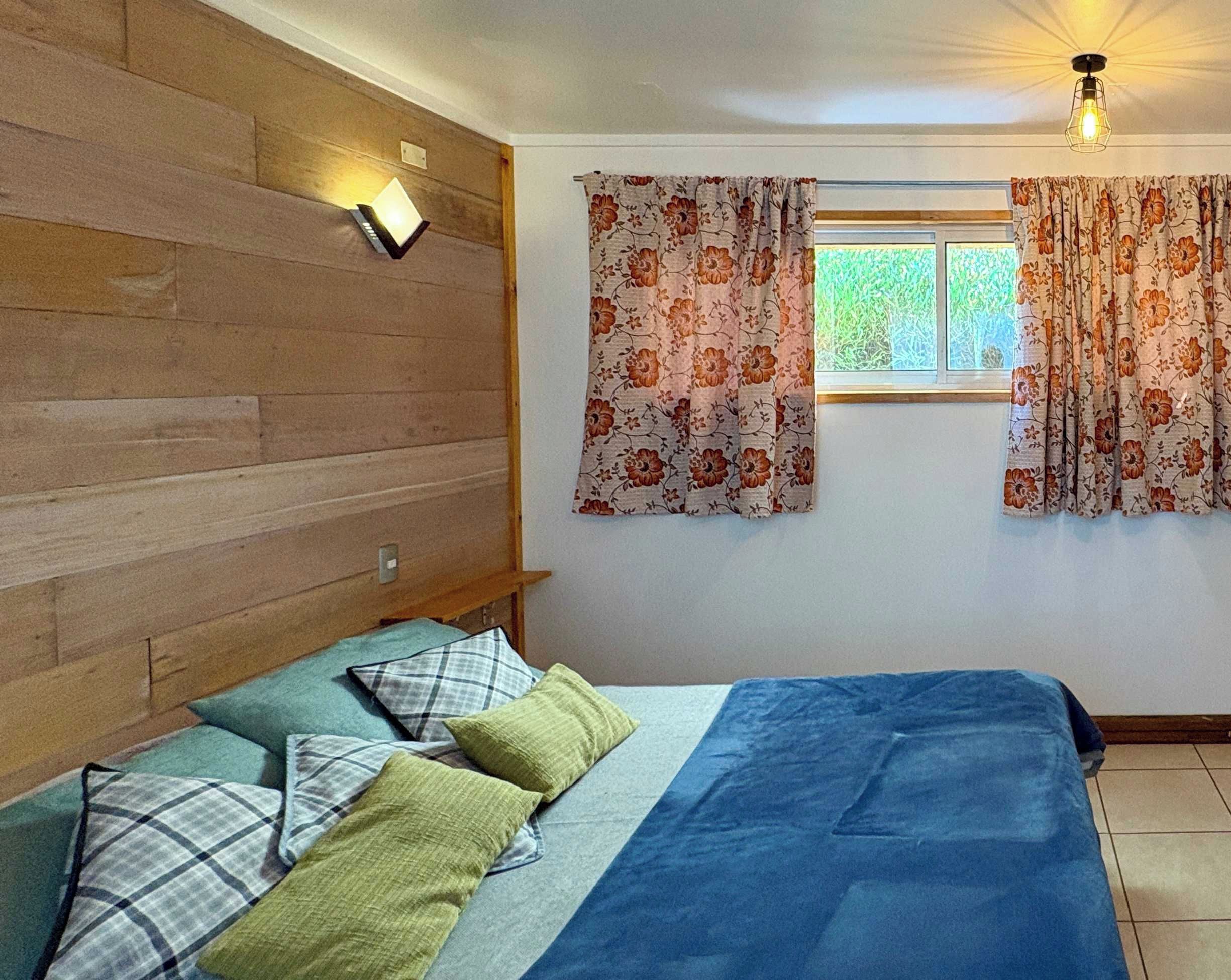Una camera da letto con un letto, cuscini, una testiera in legno, una finestra con tende e un punto luce a parete.