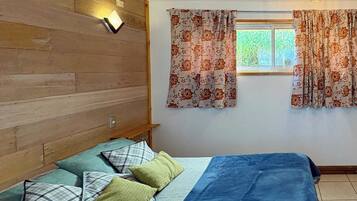 Una camera da letto con un letto, cuscini, una testiera in legno, una finestra con tende e un punto luce a parete.