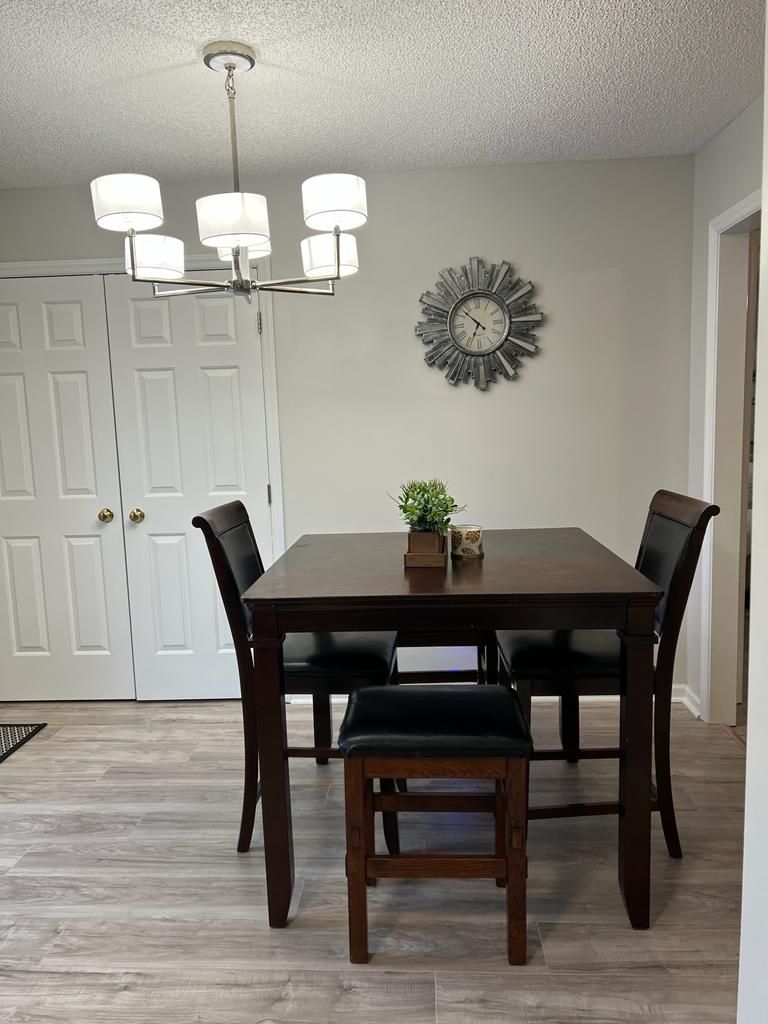 A dining room with a wooden table, four chairs, a chandelier, a clock, and a small plant on the table.
