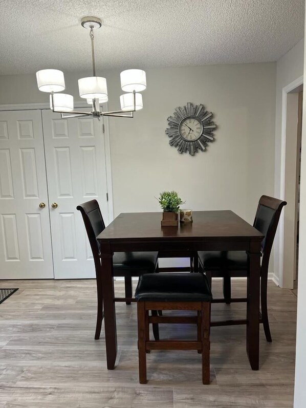 A dining room with a wooden table, four chairs, a chandelier, a clock, and a small plant on the table.
