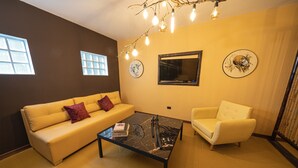 Un salon avec un canapé jaune, un fauteuil blanc, une table basse noire et un téléviseur fixé au mur.