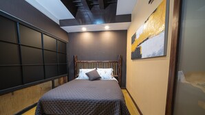 Une chambre à coucher comprenant un lit, une tête de lit, un mur orné d’un grand tableau abstrait et un plafond avec des poutres apparentes.