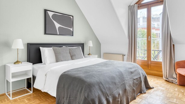 A bedroom with a bed, nightstands, a radiator, a window with curtains, and a view of buildings outside. - Historic Margareten 2BR w/ Elevator in central location, by Blueground (Vienna)