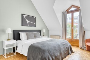 Un dormitorio con cama, mesitas de noche, un radiador, una ventana con cortinas y vistas a edificios.