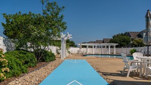 A poolside area with a blue shuffleboard court, white outdoor furniture, and a white fence.