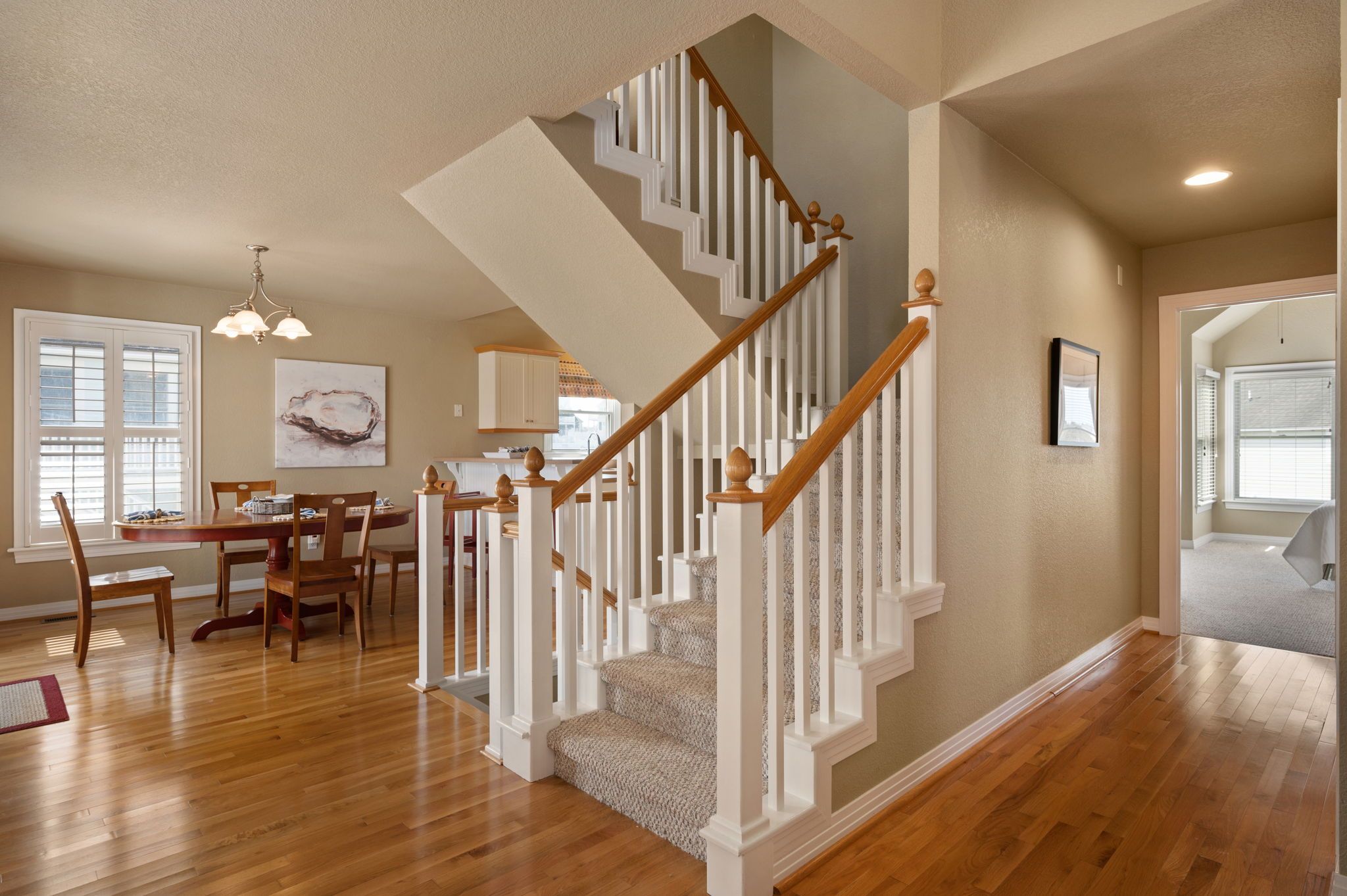 A spacious living area with a wooden staircase, a dining table with chairs, and a kitchen in the background.