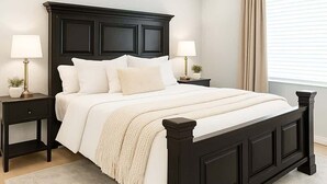 Un dormitorio con cama de madera oscura, dos mesitas de noche con lámparas, un ventilador de techo y un ventanal con cortinas.