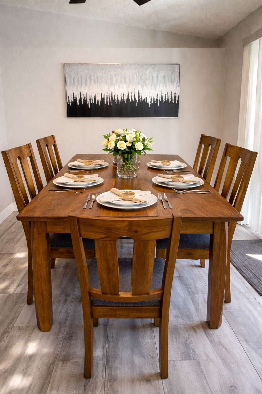 Une salle à manger avec une table en bois dressée pour six personnes, entourée de chaises en bois. Un vase rempli de fleurs blanches est placé au centre de la table.