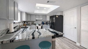 Una cocina moderna con encimera de mármol, dos taburetes de color turquesa, un refrigerador negro y un tragaluz sobre la isla.