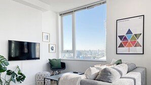 A modern living room with a large window, a flat-screen TV, and a city view. - The Designist: Downtown Apt with Water Views, Pool, Parking, AC (Vancouver)