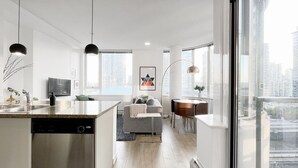 A modern kitchen with a dishwasher, a dining area with a round table and chairs, a gray sofa, and a large window offering a city view. - The Designist: Downtown Apt with Water Views, Pool, Parking, AC (Vancouver)