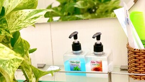A bathroom with a glass shelf holding two bottles of shampoo and foam bath, a toothbrush, and a woven basket. - Nama Hotel (Can Tho)