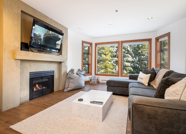 Ein modernes Wohnzimmer mit Kamin, einem großen Flachbildfernseher darüber, einem grauen Sofa mit Kissen und einem großen Fenster mit Blick auf schneebedeckte Bäume.