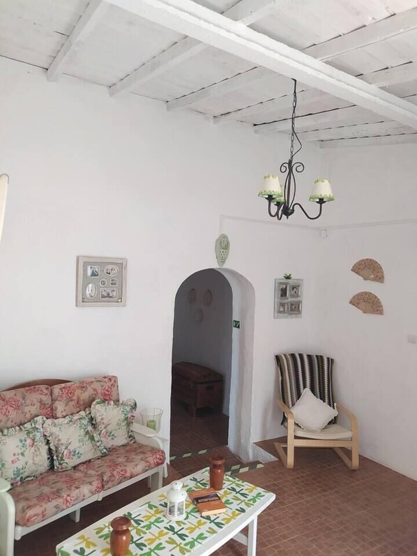 A living room with a floral sofa, a wooden chair, a table with a patterned tablecloth, and a chandelier.