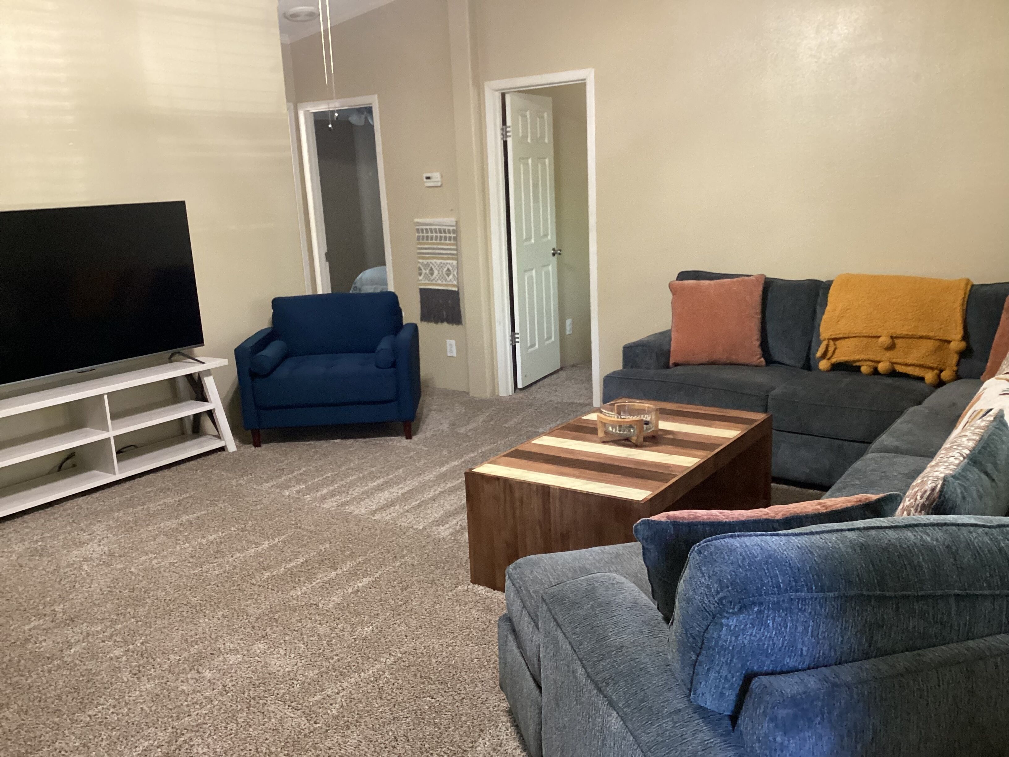 A living room with a flat-screen TV on a stand, a blue sofa, a grey sofa with a yellow throw blanket, and a coffee table with a glass candle holder.