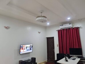 A room with a ceiling fan, a mounted TV, a desk with a computer, and a red curtain. - Smart 3 bed apartment with fast Wi-Fi & 24hr light (Abuja)