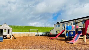 A playground with a wooden playhouse and slides, surrounded by a wooden fence and a grassy hill in the background. - 3 Bedroom Holiday Home in Newquay - Valley Views (Newquay)