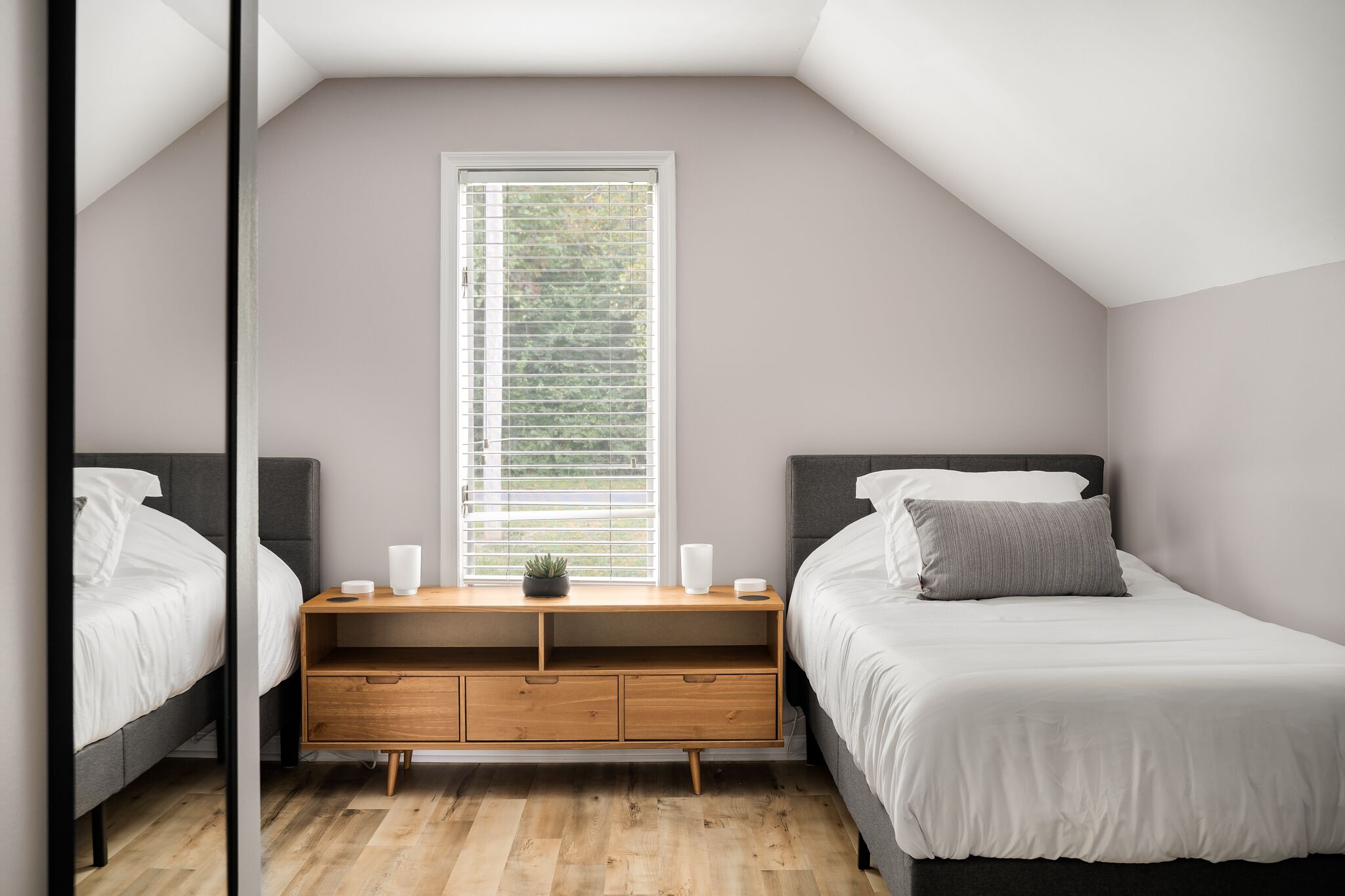 A bedroom with two beds, a wooden nightstand, and a window with blinds.