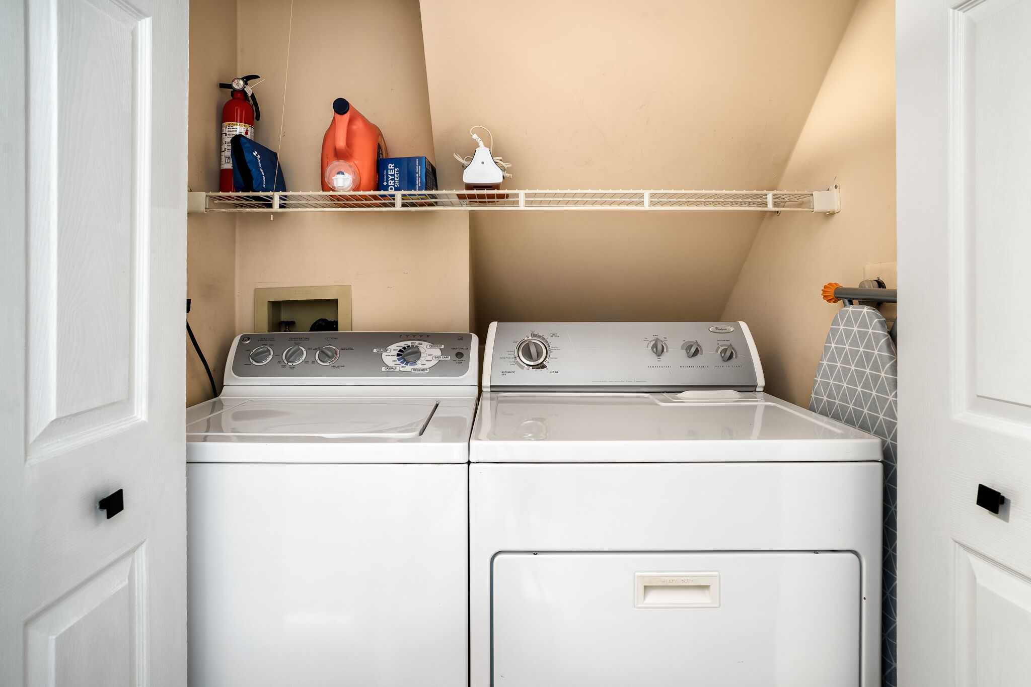 A laundry room with two white front-loading washers and dryers, a shelf with cleaning supplies, and a small ironing board.