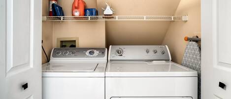 A laundry room with two white front-loading washers and dryers, a shelf with cleaning supplies, and a small ironing board.