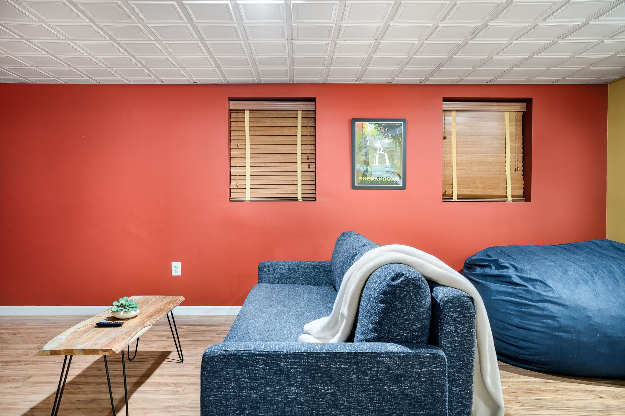 A modern living room with a red wall, two windows with blinds, a blue sofa, a wooden coffee table, and a large bean bag.