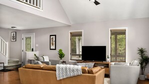 A modern living room with a sofa, armchair, and a flat-screen TV. There are windows with blinds, a staircase, and a plant. - Mountain cabin near national park & wineries (Linden)