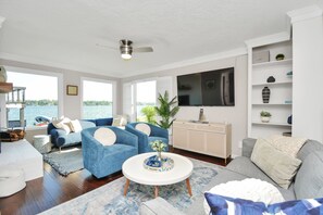 Relax in this stylish living room with a sofa, coffee table, and flat-screen TV, offering a serene view of the water through large windows.